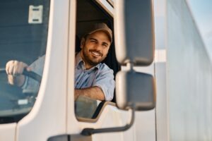 A smiling truck driver leaning out of his truck while thinking about the different types of commercial driver’s licenses in California.