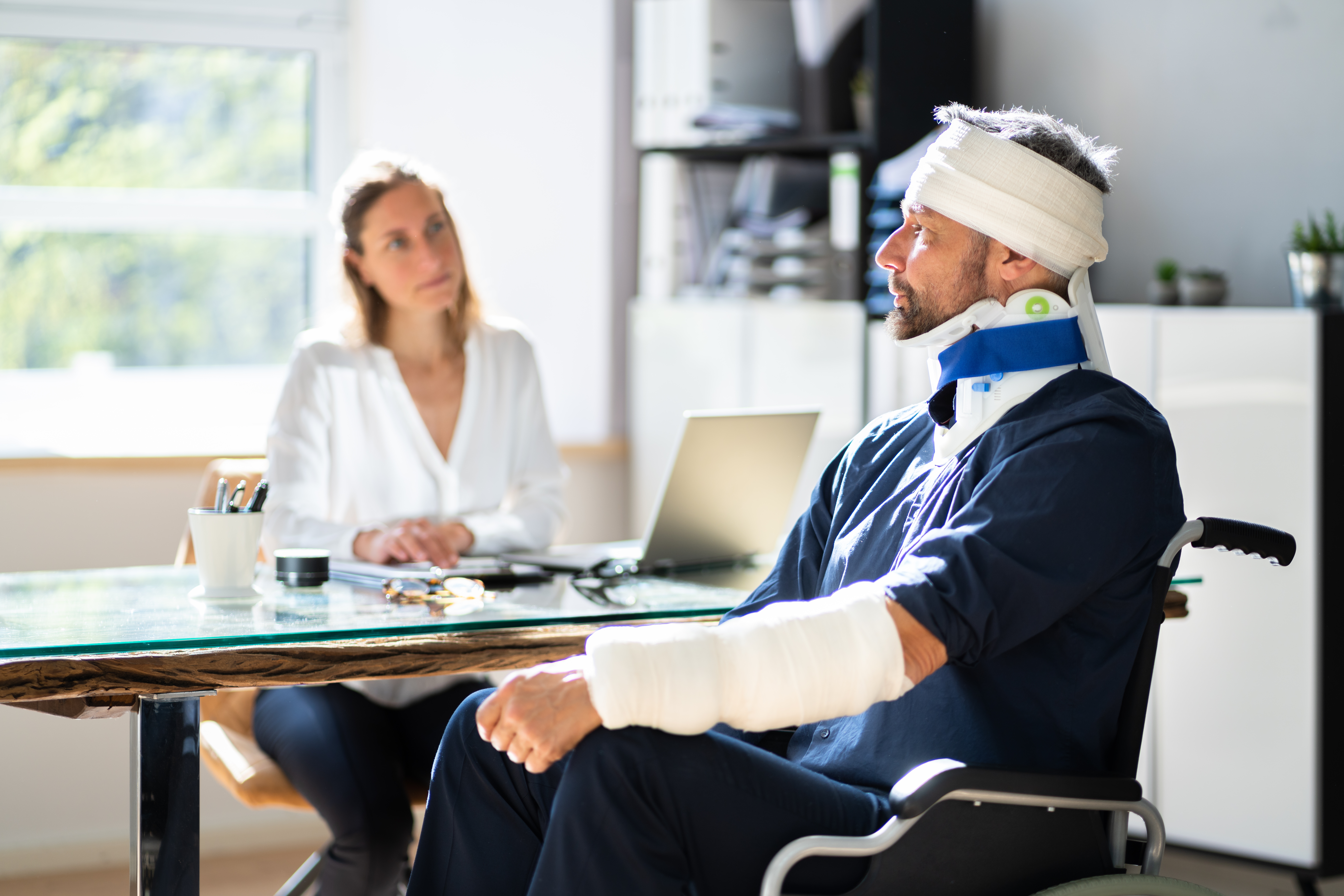A personal injury lawyer in Los Angeles meeting with an injured man in a wheelchair with a head bandage, a neck brace, and an arm cast.