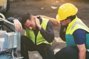 Construction worker comforting a kneeling coworker who is holding his neck in pain after a fall, which is the number-one cause of death in construction.