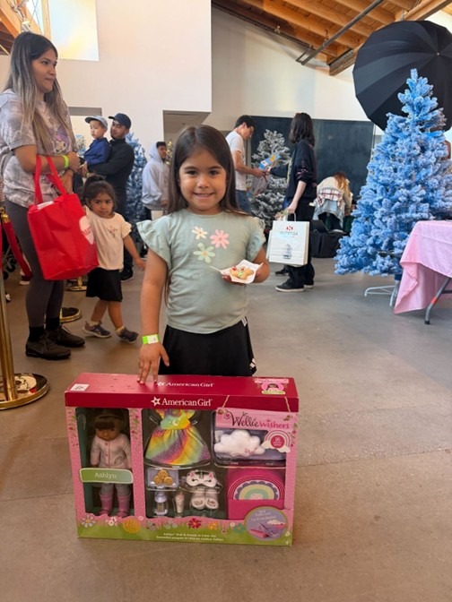 Niña pequeña sonriendo y mostrando el nuevo set de muñecas American Girl que recibió en la Campaña de Juguetes Navideños 24LA patrocinada por Omega Law Group.