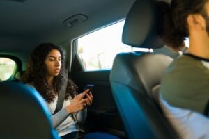 A rideshare passenger uses a cellphone while the driver steers the vehicle. Increased rideshare use after the Super Bowl can lead to accidents. An attorney can explain liability in Uber and Lyft crashes.